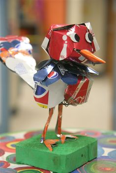 a bird made out of cans sitting on top of a green block with scissors in it's beak