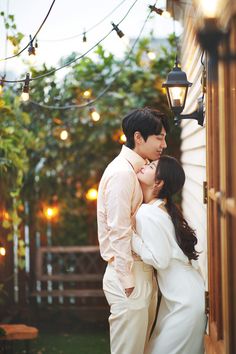 a man and woman standing next to each other in front of a house with string lights