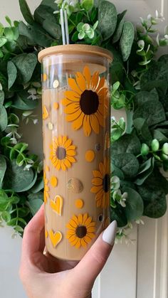 a hand holding a coffee cup with sunflowers painted on it and a green wreath behind it