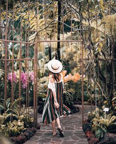 a woman wearing a white hat and striped dress walking through an archway in a garden