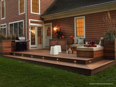 a deck with lights on it next to a house in the evening hours and an outdoor grill