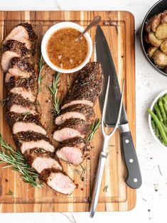 sliced pork on cutting board with side dishes and utensils next to plated vegetables