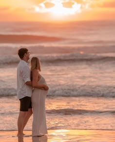 a pregnant couple standing on the beach at sunset