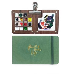 a wooden clipboard with an artist's paint set on it and a green notebook