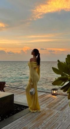 a woman in a yellow dress is standing on a dock by the ocean at sunset