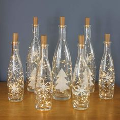 six glass bottles with lights in them on a wooden table next to a wall and a christmas tree