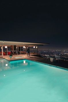 a man standing in front of a swimming pool at night with the city lights lit up