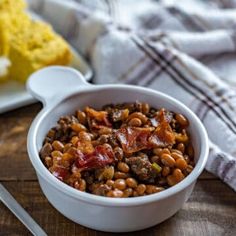 a white bowl filled with beans and meat on top of a wooden table next to bread