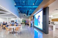 people are sitting at tables and watching a video on a large screen in the middle of a room