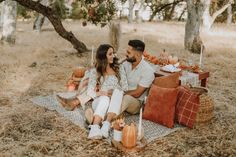 a man and woman sitting on a blanket in the woods with pumpkins around them