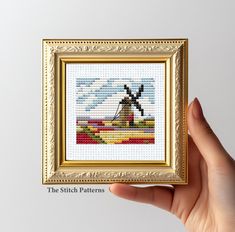 a hand holding up a cross stitch picture frame with a windmill in the middle and clouds behind it