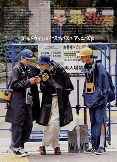 three young men standing next to each other on the sidewalk looking at their cell phones