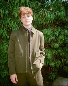 a young man standing in front of a bush wearing a green jacket and black tie