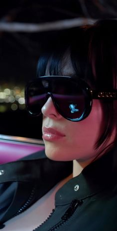 a woman wearing sunglasses sitting in the back seat of a car at night with city lights behind her