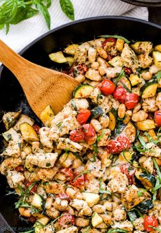 a skillet filled with chicken, zucchini and tomatoes next to a wooden spoon