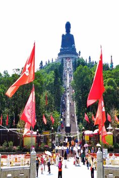 many people are standing in front of a large statue with red flags flying from it