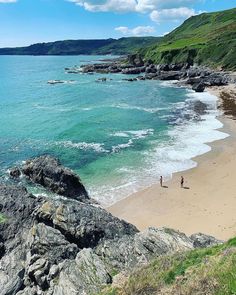 two people are walking on the beach near the water's edge, with green hills in the background