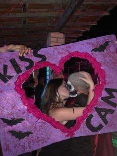 two people standing next to each other in front of a heart shaped sign that says kiss me i can