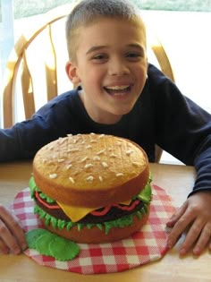 a young boy sitting at a table with a giant hamburger cake