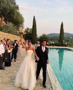 a bride and groom are walking towards the camera with their arms in the air near a pool