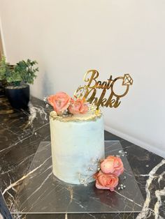 a white cake with pink flowers on top sitting on a black table next to a potted plant