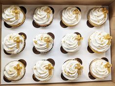 twelve cupcakes with white frosting and gold decorations in a box on a table