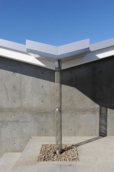 the corner of a concrete building with a metal pole and gravel in front of it