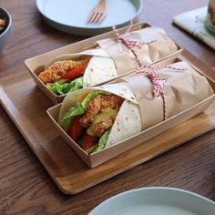 two burritos wrapped in paper on top of a wooden tray next to plates