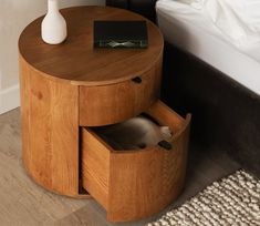 a round wooden table with two drawers and a white vase on the top, next to a bed