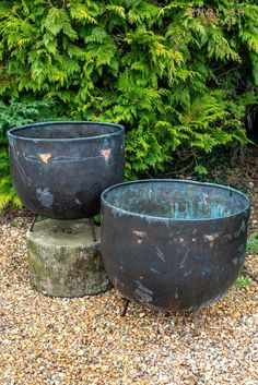 two large metal pots sitting next to each other on top of gravel covered ground with trees in the background