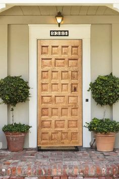 two potted plants are next to a wooden door on a brick walkway in front of a white house