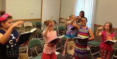 several children are standing in a room with chairs and one girl is holding an open book