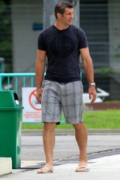 a man standing next to a green trash can on the side of the road with his hand in his pocket