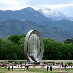 people are standing in the grass near a fountain with mountains in the background and snow - capped peaks