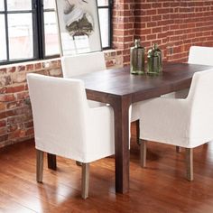 a dining room table with white chairs next to a brick wall and windows in the background