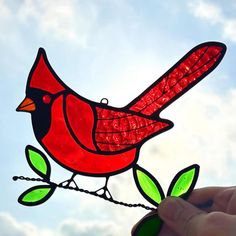 a hand holding a stained glass bird with green leaves on it's tail, against a blue cloudy sky