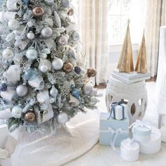 a white christmas tree with blue and silver ornaments on it, surrounded by presents in front of a window