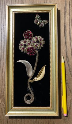 a framed painting with flowers and butterflies on it next to a yellow pencil, which is also in front of a black background