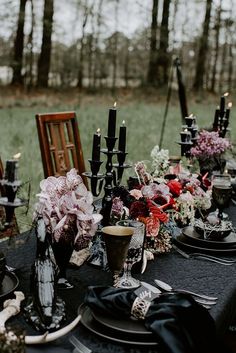 a table set up with black plates and candles for an outdoor dinner party in the woods