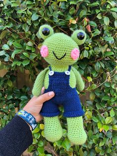 a crocheted frog stuffed animal being held by a person's hand in front of some bushes