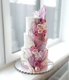 a three tiered wedding cake with pink flowers and feathers on the top, sitting in front of a window