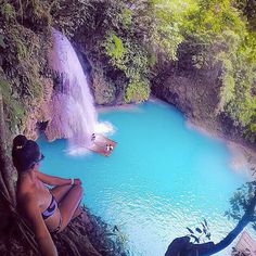 two women in bikinis sitting on the edge of a waterfall and looking at a blue pool