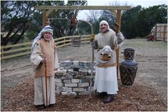 two men dressed in medieval clothing standing next to each other near a stone wall and fence