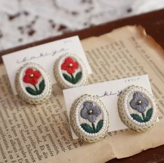 two earrings with flowers on them sitting on top of an open book next to a piece of paper