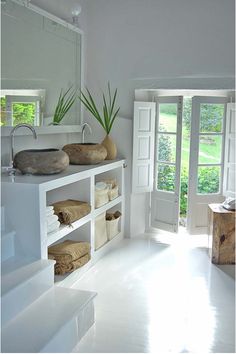 a white bathroom with two sinks and open doors leading to the outside area, while another sink sits in the middle of the room