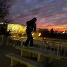 a man standing on top of a bench in front of a building at night with the sun setting behind him