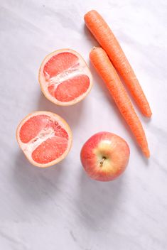 an apple, carrots, and grapefruit on a marble counter top with one cut in half