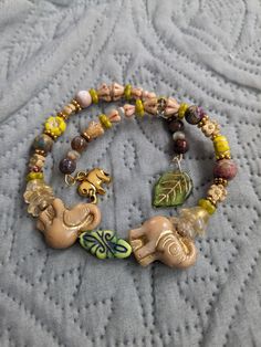 Lovely beaded charm Adjustable bracelet with two beige Chezh glass elephants and more! In the center of the elephants with raised trunks is a green Chezh glass bead with a painted black design. Chezh glass Flower beads in tan greens, and yellows. Goldstone spacer beads, and Round Jasper beads.  This is a double wrap bracelet and has two charms!  Gold tone elephant charm and a green Chezh glass leaf charm!  This bracelet is adjustable and will fit most wrists! Gift it for Mother's Day, Birthday, and more! Bohemian Beige Stretch Bracelet With Round Beads, Bohemian Beige Round Beads Stretch Bracelet, Beaded Elephant, Elephant Charm Bracelet, Bead Flowers, Double Wrap Bracelet, Elephant Charm, Flower Beads, Leaf Charms