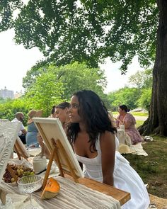 a woman sitting in front of an easel with food and wine on the table