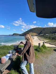 a woman is cooking on the side of a road by the beach and water with mountains in the background